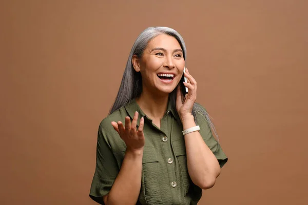 Mature lady has pleasant phone conversation. Cheerful smiling senior grey-haired business woman talking on smartphone standing isolated on brown — ストック写真