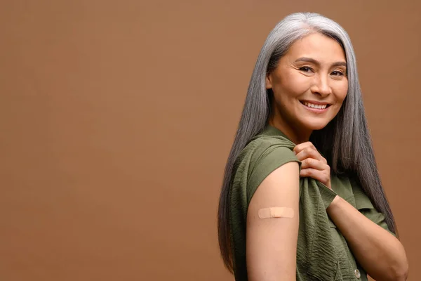 Smiling senior grey-haired woman with a medical patch after vaccination isolated on brown background, protecting hand with bandage after injection
