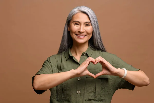 Love you. Portrait of attractive romantic mature woman standing and making heart with hands, while smiling playfully. Indoor studio shot isolated on brown — ストック写真