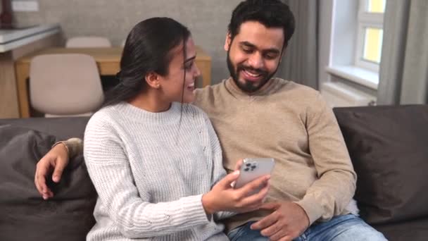 ソーシャルネットワークで余暇を過ごすソファの上にスマートフォンを持つインドのカップル — ストック動画