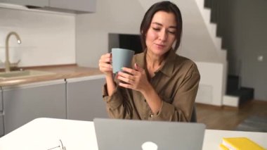 Rahat Asyalı çok ırklı kadın serbest çalışan dizüstü bilgisayarın önünde bir fincan kahveyle rahatlıyor.