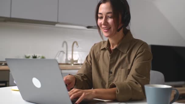 Sorrindo jovem funcionária vestindo fone de ouvido sem fio sentado com computador na mesa — Vídeo de Stock