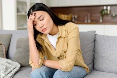 Stresli genç Asyalı kadın kafasına dokunuyor ve başı dönüyor, baş ağrısı ve migren ağrısı çekiyor, evdeki kanepede oturuyor.