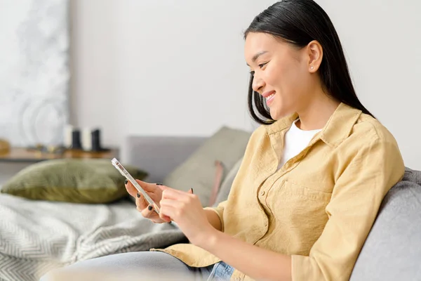 Oldalnézet a nyugodt ázsiai nő ül a kanapén a hangulatos nappaliban, és gazdaság okostelefon. Nő időt töltenek online, SMS-t a barátaival — Stock Fotó