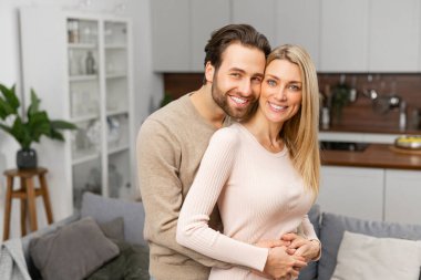 Happy married couple posing for a photo on kitchen. Happiest hearts make the happiest homes, caucasian husband hugging his wife from behind