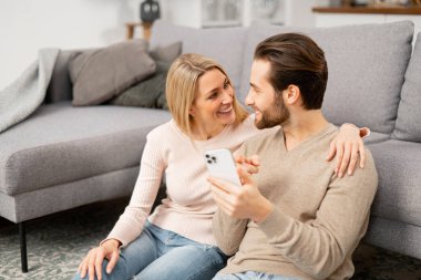 Gülümseyen beyaz çift evdeki rahat kanepenin yanında otururken akıllı telefon kullanıyor. Neşeli eşler cep telefonu kullanıyor, film izliyor.