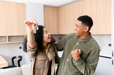 Multiracial woman holding home keys while hugging boyfriend in new apartment. Lovely couple feeling ecstatic about their recent purchase