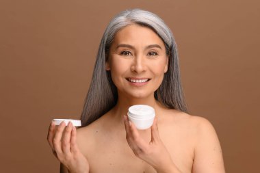 Serene orta yaşlı, sağlıklı ciltli Asyalı kadın bir kavanoz nemlendirici anti-yaşlanma kremiyle ayakta duruyor.