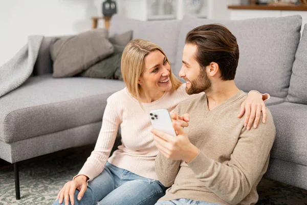 Gülümseyen beyaz çift evdeki rahat kanepenin yanında otururken akıllı telefon kullanıyor. Neşeli eşler cep telefonu kullanıyor, film izliyor.