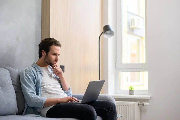 Concentrated caucasian guy looking for issues solving, businessman sitting at the sofa and staring thoughtfully at the laptop screen — Fotografia de Stock