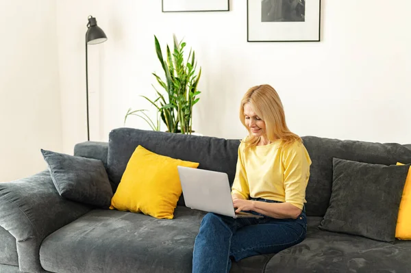 Mutlu yaşlı kadın oturma odasında koltukta rahatça oturur, dizüstü bilgisayarda çalışır, gülümser. Modern olgun 60 'lı yaşlardaki kadınlar hafta sonlarını evde geçirip kablosuz internete bakıyorlar.