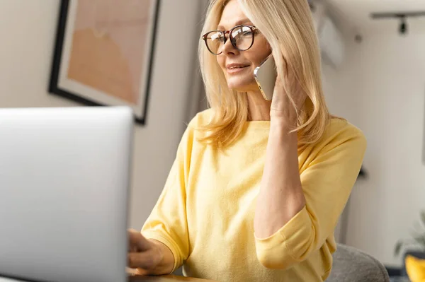 Sorrindo tipo mulher sênior trabalha em casa. Ela fala ao telefone enquanto se senta na mesa em frente ao laptop — Fotografia de Stock