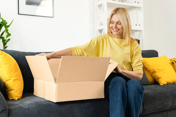 Happy senior mature adult woman is looking at her new parcels, sitting on the couch, and unpacking. Smiling online lady opening online store order