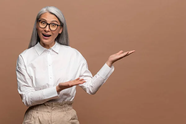 Portrait of surprised mature woman pointing finger and palm right at empty space, looking at camera with open mouth and shocked expression. Indoor studio shot isolated on beige — Foto de Stock
