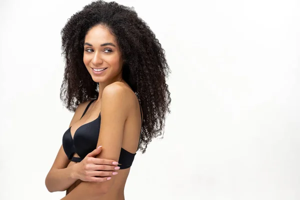 Love and care yourself. Delightfully charming and attractive young woman with curly hair hug herself and look away over white background, African-American female in black bra — Stock fotografie