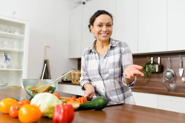 Neşeli genç kadın yemek pişirme hakkındaki video blogunu kaydediyor. Kadın salata hazırlar, sebze doğrar ve kameraya bakar.