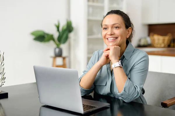 Smiling business woman using laptop for remote work from home. Confident lady checking emails, conducting business correspondence while sitting at the table