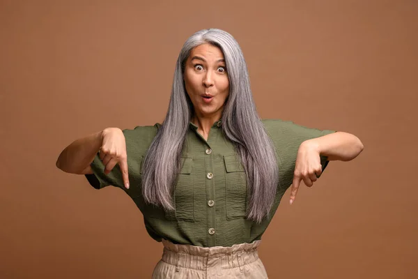 Cheerful positive woman pointing fingers down showing space for your advertisement, looking at camera with surprised face. Indoor studio shot isolated on brown — Stock Photo, Image