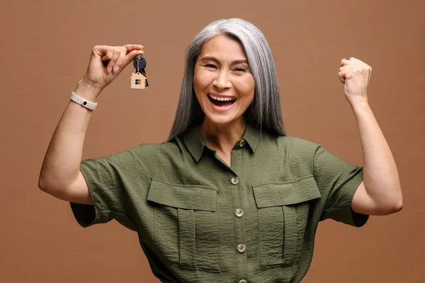 Happy overjoyed senior woman holding the keys with keychain in form of little house isolated on brown background. Smiling woman holding keys from new property, happy buyer of own estate — ストック写真