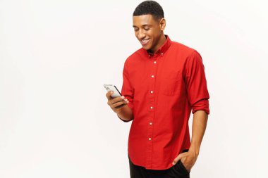 Smiling optimistic African-American young man in red casual shirt using smartphone isolated on white, multiracial freelancer or student gay chatting online, texting, messaging, web surfing