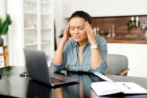 Frustrated young woman using laptop computer suffering from headache, sitting in home office and holding head with hands, feels sick and unwell — Fotografia de Stock
