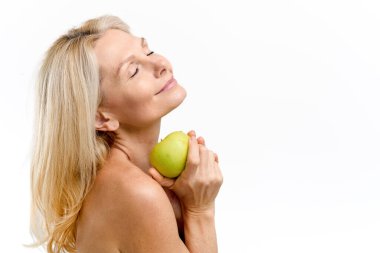 Orta yaşlı, çıplak omuzlu, gözleri kapalı poz veren, beyaz tenli bir model. Mutlu, olgun, 50 'li yaşlarda, elinde taze yeşil bir elma tutan ve gülümseyen bir kadın.