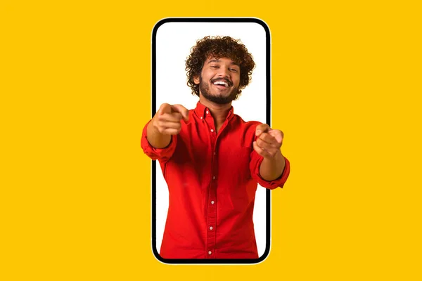 Handsome curly Indian guy looking out from huge smartphone and points fingers at camera — Stockfoto