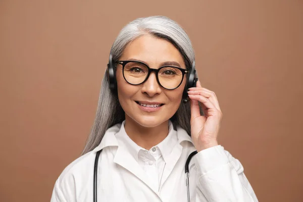 Middle-aged Asian woman doctor in white medical gown with stethoscope using wireless headset — ストック写真