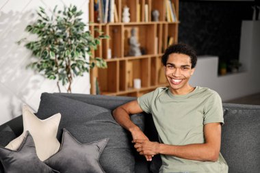 Ethnic man sitting on the sofa and smiling, in a normal t shirt, feeling comfort and relaxation, at an interview