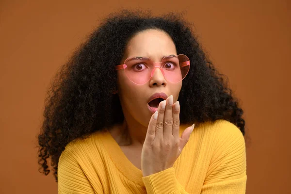 Shocked and amazed young attractive woman looks at the camera isolated on brown, a curly multiracial girl screams surprised, holding hands near her face — Stock Photo, Image