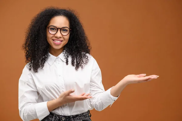 Charmig multiracial kvinna med afro frisyr indikerar handflator på tomt utrymme isolerad på brun bakgrund, leende kvinnliga student reklam — Stockfoto