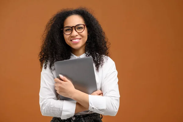 Mladá Afroameričanka s afro účesem v elegantním neformálním oblečení a stylovými brýlemi, stojící izolovaně na hnědé, s přenosným počítačem, s úsměvem, zaměstnankyně kanceláře — Stock fotografie