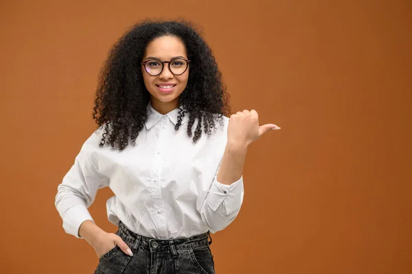 Charmig multiracial kvinna med afro frisyr som indikerar fingrar på tomt utrymme isolerad på brun bakgrund, leende kvinnliga student reklam — Stockfoto