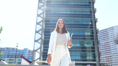 Intelligent confident businesswoman in smart casual attire with laptop on the city street, female entrepreneur in white blazer carrying laptop looking at the camera, urban landscape
