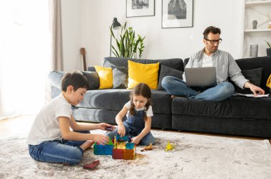 Babası dizüstü bilgisayarını evdeki kanepede rahat rahat otururken iki çocuğu yerde oynarken serbest çalışmak için kullanıyor. Meşgul bekar ebeveyn, uzakta bebek bakıcılığı yapıyor.