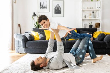 Mutlu baba ve okul çağındaki oğlu evde birlikte vakit geçiriyorlar, rahat bir oturma odasında yerde uzanıyorlar, baba çocuğu taşıyor, oğul kollarını açıyor, uçak numarası yapıyor. Sevgili baba, oğlunla oyna.
