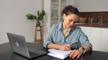 Gülümseyen bir kadın web siteleri, dersler izlemek için dizüstü bilgisayar kullanıyor, evde masa başında oturuyor ve notlar alıyor, çevrimiçi eğitim kursuna katılan bir kadın, konu araştırması yapıyor.