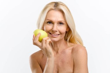 Sağlıklı ve orta yaşlı, çıplak omuzlu, yüzünün yanında yeşil elma tutan, kameraya bakan ve geniş gülümseyen güzel bir kadın.