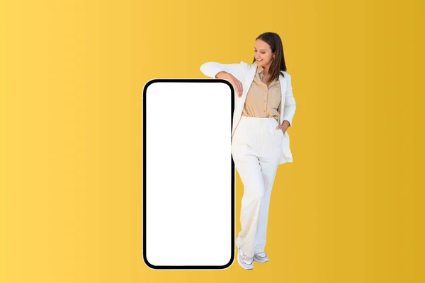 Best deal promotion. Smiling positive business lady leaning on huge smartphone with empty blank screen, looking at it, female office employee recommending new app or website, mockup