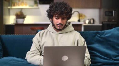 Hayal kırıklığına uğramış kıvırcık Hindistanlı serbest çalışan adam ofisteki kanepede otururken bir hata yaptı, çok ırklı erkek öğrenciyi üzdü ve umutsuzluk içinde laptop ekranına baktı.