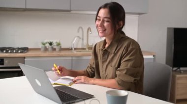 Female student watching a lecture, webinar using a laptop, sits at the table and writes with a pen in a notepad. Asian young woman employee takes a note watching online training, working remotely