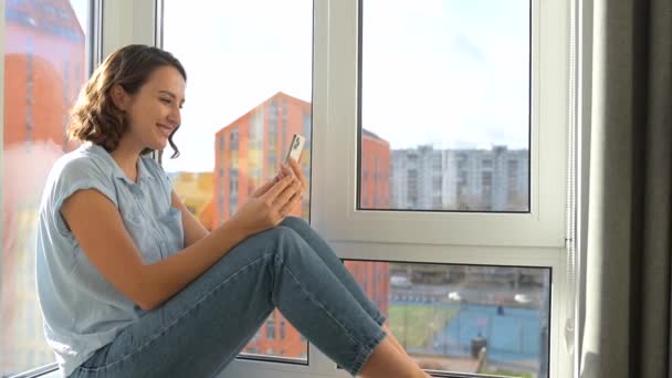窓辺に座っている間 スマホを使ってビデオ接続をしている白人女性 笑顔の女の子手を振っこんにちは 挨拶の友人や家族 ビデオ通話のためのモバイルアプリを使用して — ストック動画