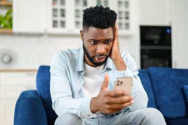 Akıllı telefon tutan, üzgün esmer genç adam, telefon ekranında hayal kırıklığı yüz ifadesiyle bakan, kötü bir yorum aldı sosyal ağlar nefret, nefret, kanepede otururken