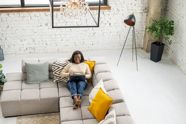 High angle view of the cheerful lady spend leisure time in network, web surfing. Relaxed multiracial woman sitting on the couch and chatting online on the laptop, answering email