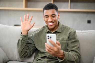 Heyecanlı, neşeli, dişlek gülümseyen ve video bağlantısı için akıllı telefonu kullanan adam. Siyah adam cep telefonuyla el sallıyor, uzaktaki koltukta konuşuyor.