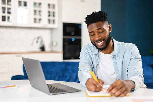 Modern bearded man working with laptop in home office. Multiracial male in smart casual shirt watching webinar and writing down, taking notes with a pen, planning schedule