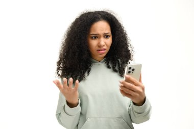 Disappointed young African-American woman holding smartphone and watching at the phone screen, lady received bad notification or messages, feels upset, shrugging, isolated