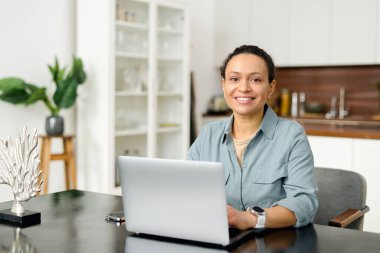 Normal giyinen kadın çalışan kameraya bakar ve gülümser, mesajlar yazar, e-postaları cevaplar, ön görüş. Kendine güveni tam orta yaşlı bir kadın modern ofiste dizüstü bilgisayarla çalışıyor.