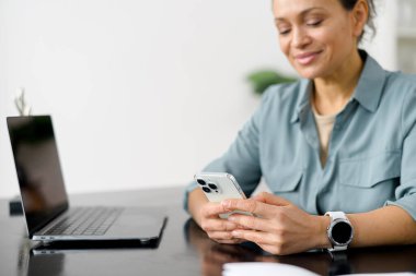 Kıvırcık saçlı, neşeli, beyaz bir kadın masada oturuyor ve akıllı telefon kullanıyor. İnternette güzel bir bayan ofis çalışanı telefonda konuşuyor.