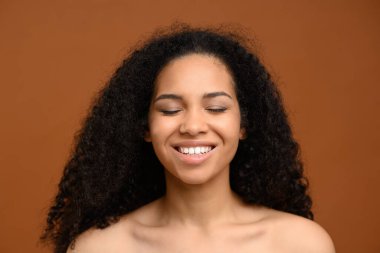 Kahverengi duvarda duran çok kültürlü kadının portresi. Genç ve güzel Afro-Amerikalı bir kadın kahverengi arka planda kapalı gözlerle poz veriyor ve dişlek gülümsüyor. Doğal güzellik konsepti